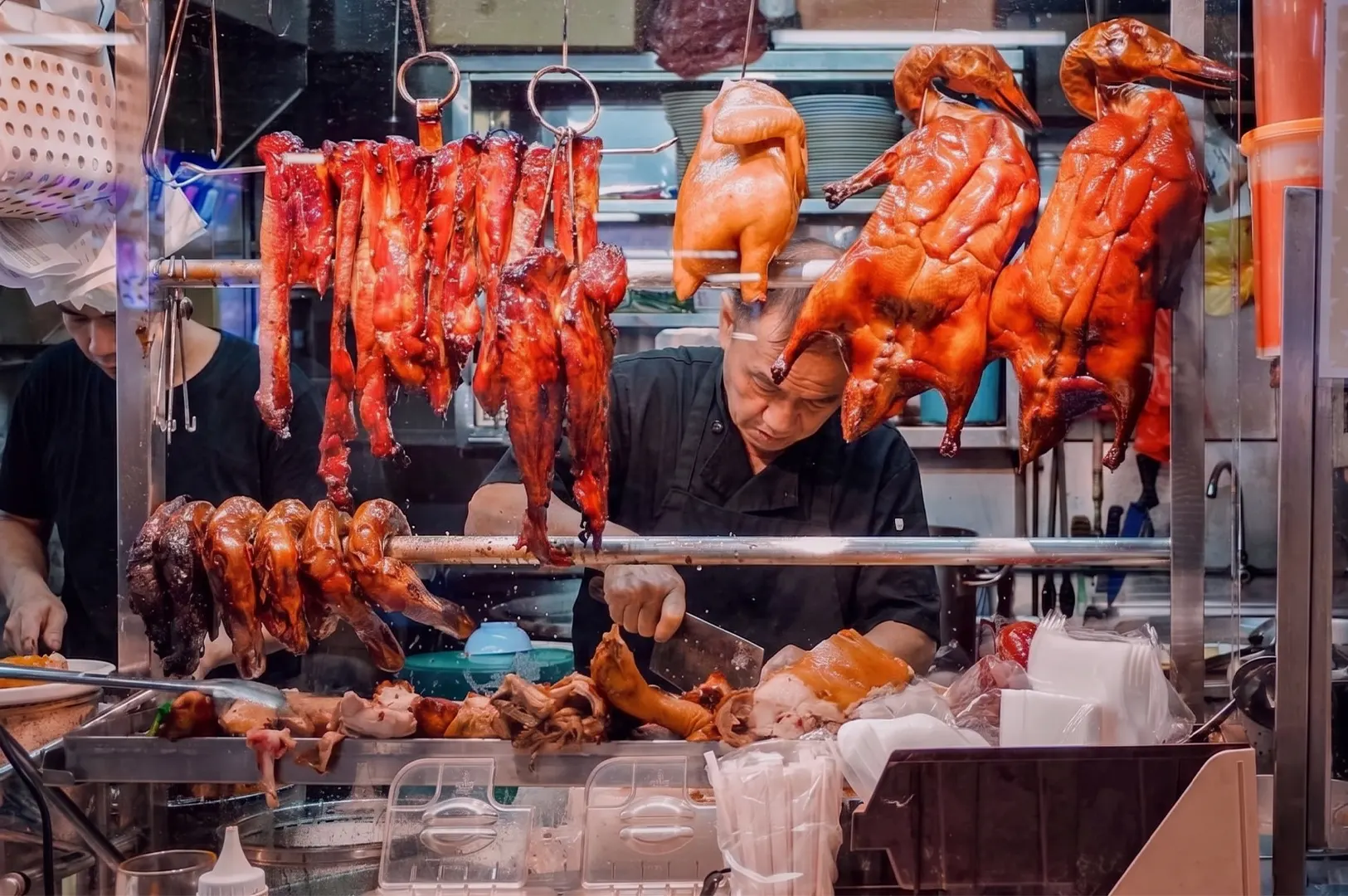 A traditional street food vendor chopping meat behind a glass display of hanging roasted ducks and pork at an Asian night market stall.